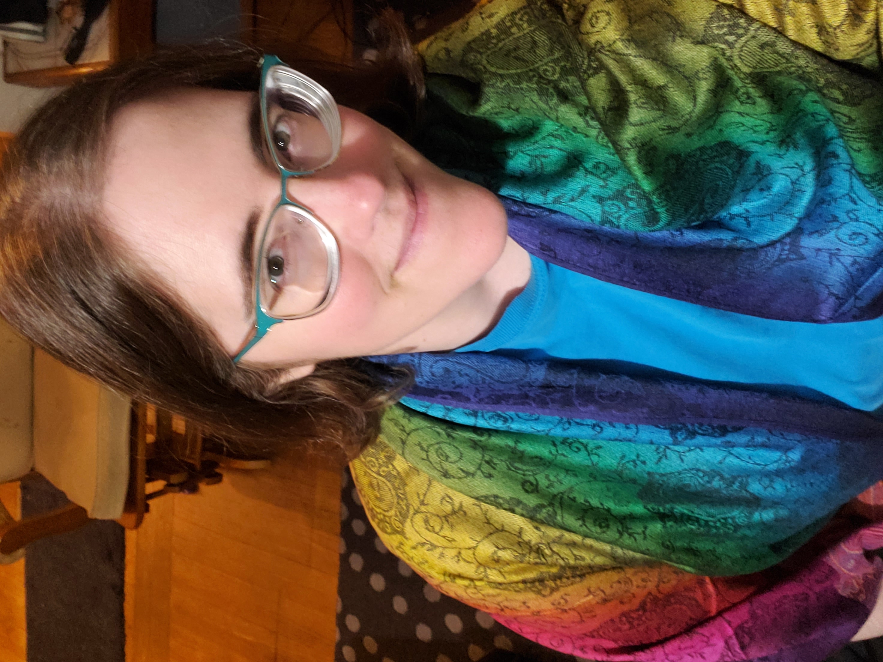 A selfie of the author. A white woman with medium brown hair and glasses looks up towards the viewer with a playful smile. She is wearing a bright blue shirt and is wrapped in a bright rainbow pashmina scarf. 