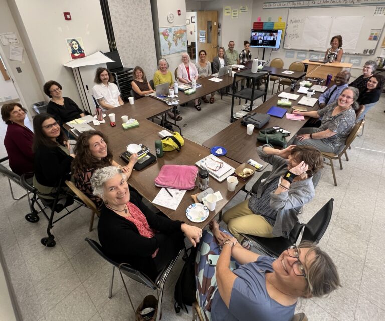 Staff smiling around a classroom table, with additional staff remotely on a monitor.